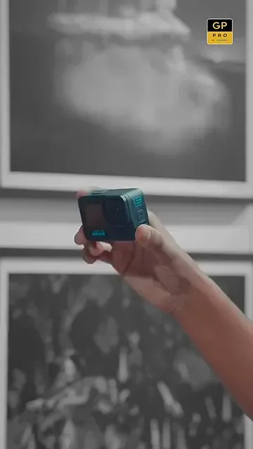 Mão segurando uma câmera de ação diante de uma fotografia em preto e branco numa parede de galeria.