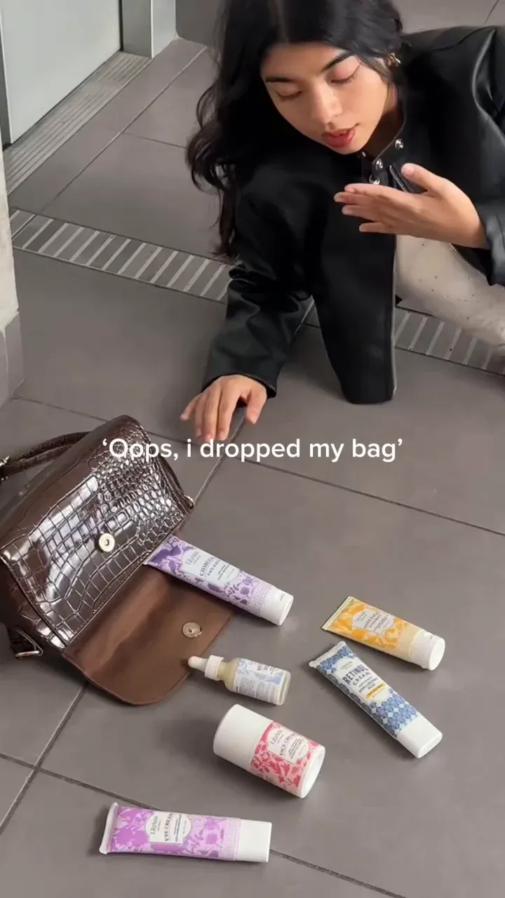 Modelo agachada ao lado de uma bolsa caída, com os produtos de beleza espalhados pelo chão.
